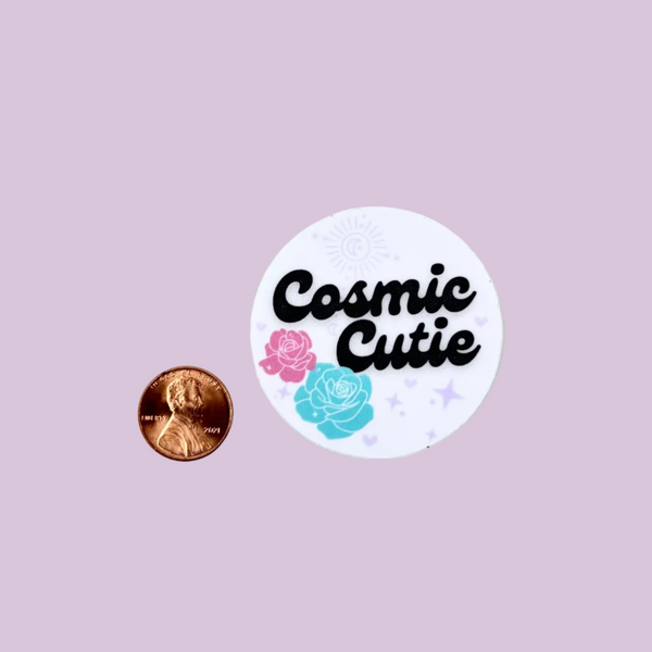 Round sticker with 'Cosmic Cutie' text and flowers next to a penny for scale on a light pink background